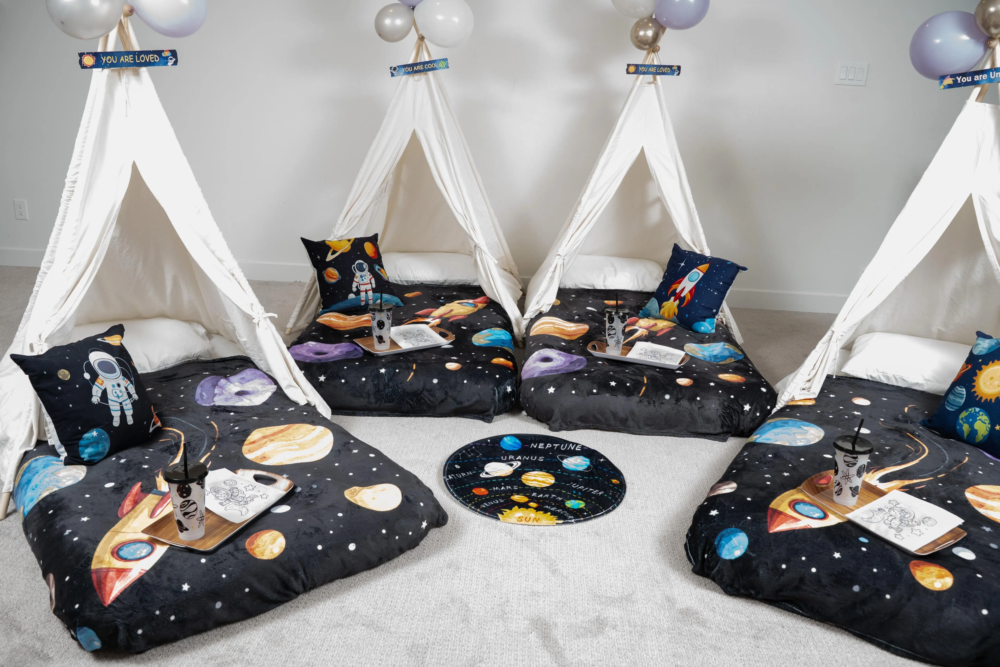Several white teepees with space-print bedding, astronaut pillows, balloons and trays.